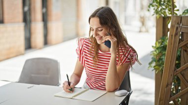 Kafe terasında akıllı telefondan konuşan genç sarışın kadın deftere yazı yazıyor.