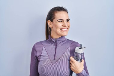 Spor kıyafetleri giymiş güzel bir kadın elinde su şişesi, yüzünde gülümsemeyle yan yana bakıyor, doğal bir ifade. Kendinden emin gülen. 