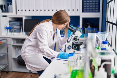 Young caucasian woman scientist using microscope write on document at laboratory