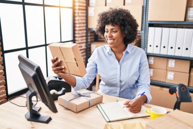 African american woman ecommerce business worker writing on notebook holding package at office