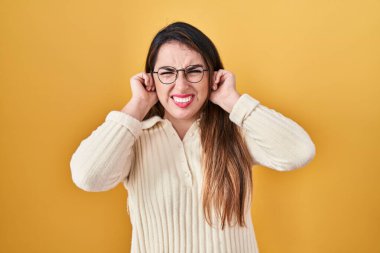 Young hispanic woman standing over yellow background covering ears with fingers with annoyed expression for the noise of loud music. deaf concept. 