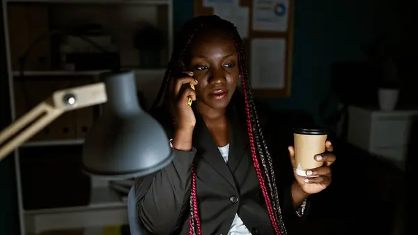 Serious african american woman boss striding towards success, engrossed in deep work conversation via smartphone, sipping espresso amidst dark office ambiance