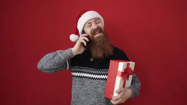 Young Redhead Man Holding Christmas Gift Talking Smartphone Isolated Red — Stock Photo, Image