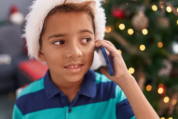 自宅でクリスマスツリーで立っているスマートフォンで話す愛らしいヒスパニックの少年 — ストック写真