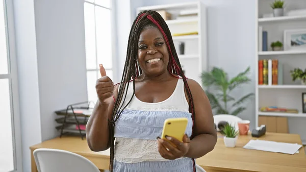 Smiling african american woman boss gives a thumb-up gesture while working on her phone in the office, embodying elegance and success in the midst of her professional duties