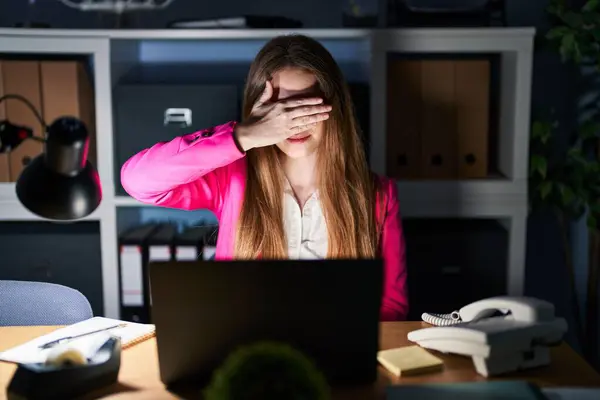 Young caucasian woman working at the office at night smiling and laughing with hand on face covering eyes for surprise. blind concept.