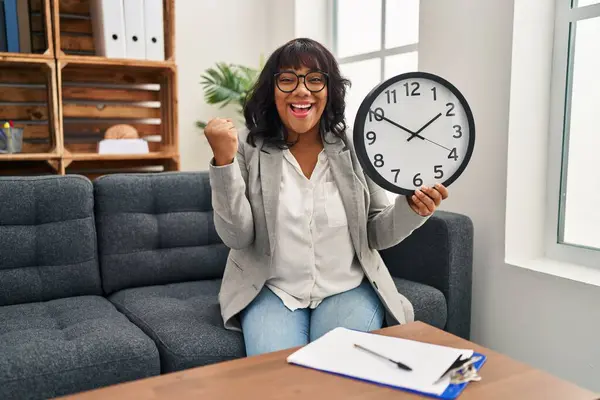 Hispanic woman working at therapy office holding clock screaming proud, celebrating victory and success very excited with raised arms