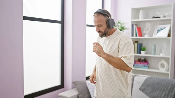 Attractive young guy, headphones on, seriously listening to music, engrossed in sound; dancing relaxedly within the intimate interior of his living room home, the expression on his face concentrated