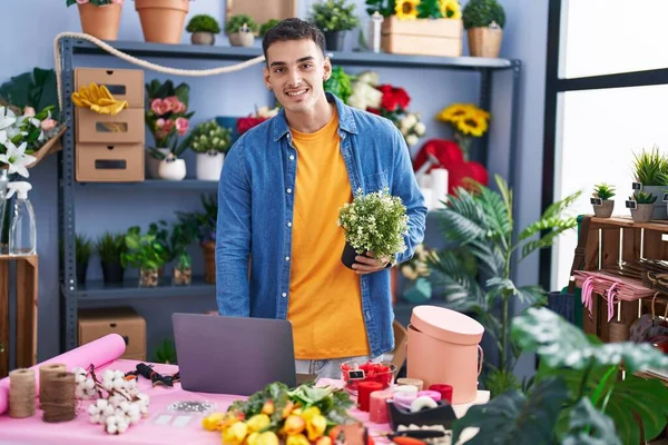 Young hispanic man florist using laptop holding plant at florist store