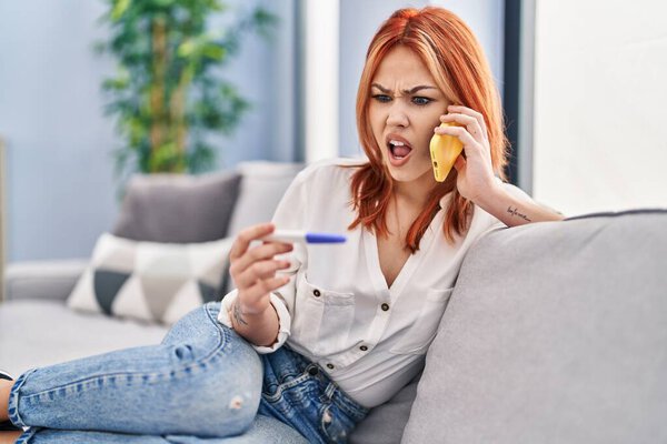 Young caucasian woman holding pregnancy test result speaking on the phone angry and mad screaming frustrated and furious, shouting with anger. rage and aggressive concept. 