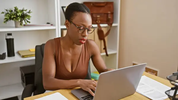 Hardworking african american woman boss, beautiful and serious, working online on laptop at office table with focused face