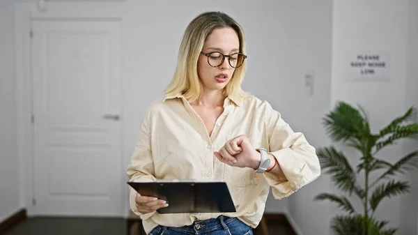 Young blonde woman business worker holding clipboard looking watch at the office waiting room