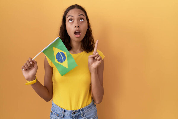 Young hispanic woman holding brazil flag amazed and surprised looking up and pointing with fingers and raised arms. 