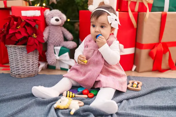 自宅でクリスマスツリーで床に座っているフープのおもちゃを吸う愛らしいヒスパニックの赤ちゃん — ストック写真