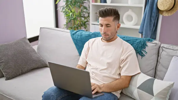 焦点を当てた若いヒスパニック系男性は オンラインで作業しながらノートパソコンに魅了され 明るく照らされた部屋の自宅の居心地の良いソファーに座って 魅力的な集中のオーラを表示します — ストック写真