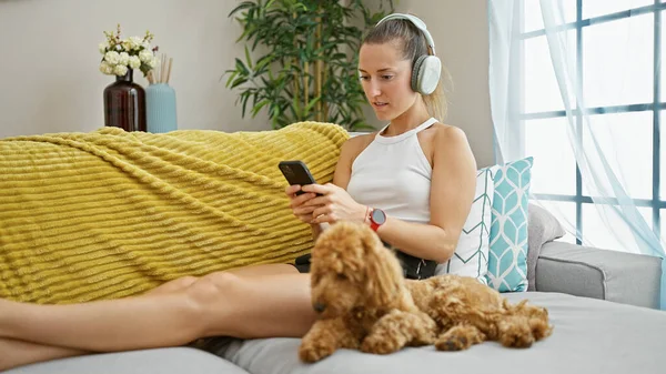 自宅でヘッドフォンを身に着けているスマートフォンを使用してソファに座っている犬を持つ若い白人女性 — ストック写真