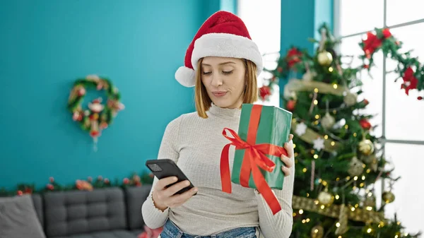Νεαρή Ξανθιά Γυναίκα Κρατώντας Χριστουγεννιάτικο Δώρο Χρησιμοποιώντας Smartphone Στο Σπίτι — Φωτογραφία Αρχείου