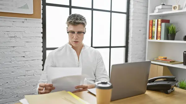 Serious-faced young caucasian male executive, engrossed in reading a business document, diligent worker thriving at his desk job in the indoor office environment.