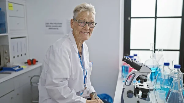 自信を持って 笑顔の灰色の髪のシニア女性科学者 彼女の仕事に満足して ラボで顕微鏡を使用して 医学研究センターで彼女の成熟した知恵に科学を提出 — ストック写真