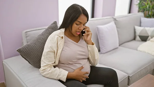 Emotional young latina expecting mother, casually touching belly on living room sofa, has a serious talk on her smartphone as water breaks in her cozy apartment home.