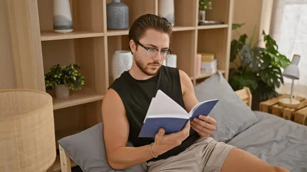 Mladý Hispánec Nosí Brýle Čtení Knihy Sedí Posteli Ložnici — Stock fotografie