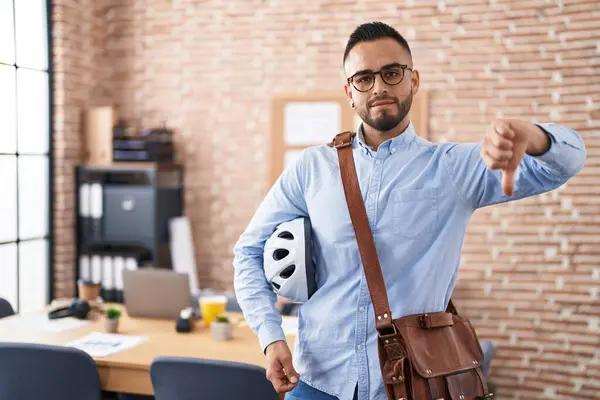 オフィスで働く若いヒスパニック男性は 怒った顔で自転車のヘルメットを保持しています 否定的なサインは 親指の下に嫌いを示しています 拒絶概念 — ストック写真