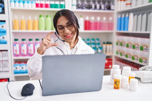 Young arab woman working at pharmacy drugstore using laptop smiling and confident gesturing with hand doing small size sign with fingers looking and the camera. measure concept.