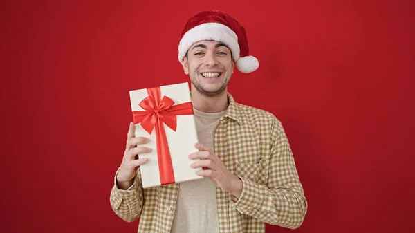 若いヒスパニック男性は クリスマスの帽子を着て自信を持って微笑み 孤立した赤い背景の上に贈り物を保持します — ストック写真