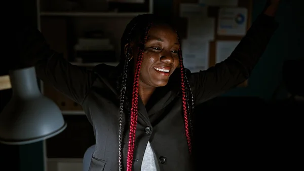 Confident african american woman boss with braids celebrating her business success, while working on a computer in a beautiful office room lit by night