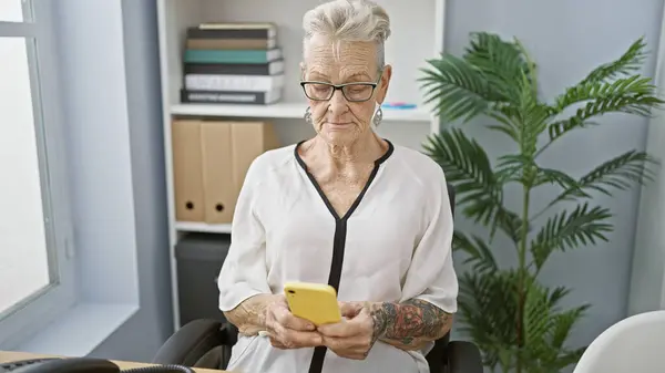 オフィスの喧騒の中でスマートフォンに真剣にメッセージを入力する 優雅で灰色の髪のシニア女性の上司 年齢と技術の絶妙なブレンド — ストック写真