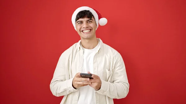孤立した赤い背景の上にスマートフォンを使用してクリスマスの帽子をかぶっている若いヒスパニック男性 — ストック写真