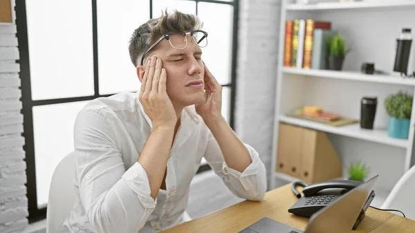 Worried young caucasian man suffering a headache at work, took off glasses, sitting in office with laptop