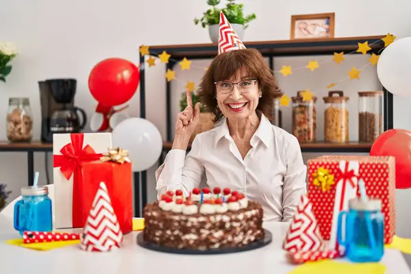 Middle age woman celebrating birthday holding big chocolate cake surprised with an idea or question pointing finger with happy face, number one
