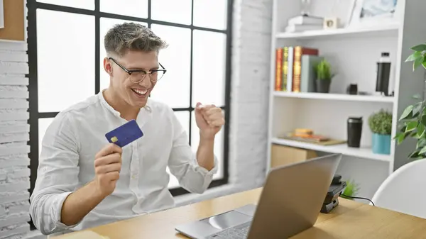Handsome young caucasian man celebrating success, shopping online with laptop and credit card at office workplace
