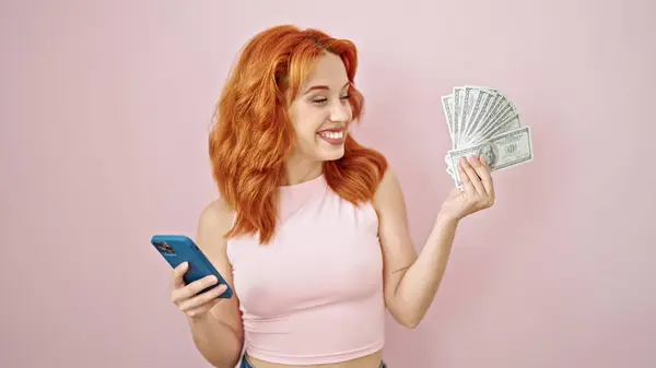 Young redhead woman smiling holding dollars and smartphone over isolated pink background