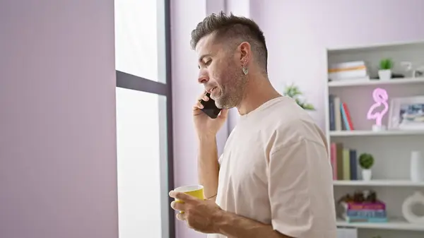 Serious young hispanic man in a deep conversation over the phone, throwing sad looks through the window while sipping coffee at home in the morning.