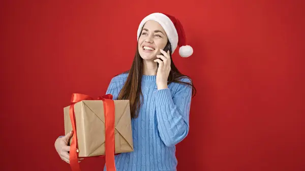年轻的高加索女人头戴圣诞礼帽 拿着礼物在智能手机上与外界隔绝的红色背景下交谈 — 图库照片