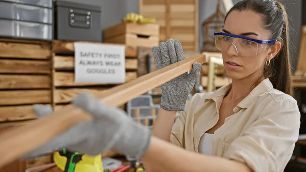Beautiful young hispanic woman carpenter skillfully examining wood plank in carpentry workshop, sporting protective glasses in a relaxed demeanor