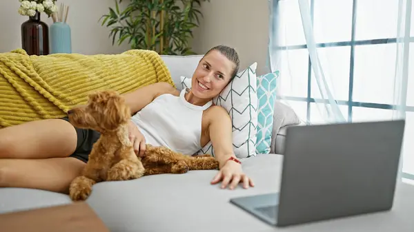 Young caucasian woman with dog watching movie on laptop lying on sofa smiling at home