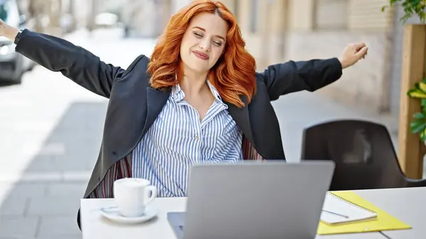Young redhead woman business worker using laptop stretching arms at coffee shop terrace