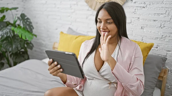 Morning glow, young, smiling, pregnant woman saying hello with her hand during a relaxed video call on her touchpad, from the cosy comfort of her bedroom bed.