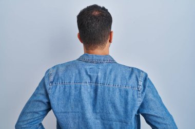 Mavi arka planda dikilen İspanyol genç adam kolları vücudunda geriye bakıyor. 