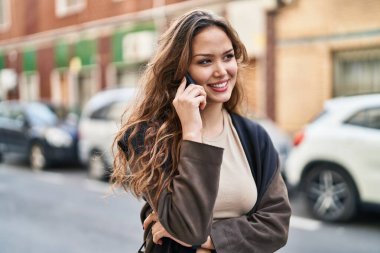 Genç, güzel, İspanyol bir kadın gülümsüyor ve caddede akıllı telefondan konuşuyor.