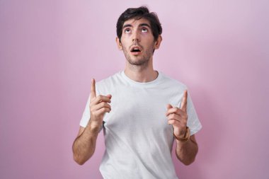 Young hispanic man standing over pink background amazed and surprised looking up and pointing with fingers and raised arms. 