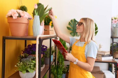 Young blonde woman florist reading document touching plant at flower shop