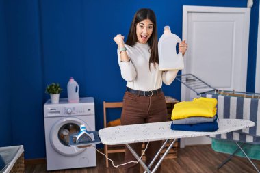 Çamaşırlarını katlamış, gururla ütü yapan genç esmer kadın zaferi kutluyor ve kollarını kaldırarak çok heyecanlanıyor. 