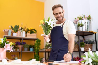 Middle age man florist holding bouquet of flowers writing on notebook at flower shop