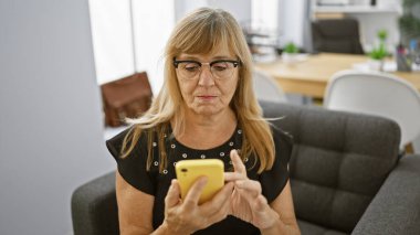 Başarılı bir iş, orta yaşlı sarışın kadın işçi akıllı telefonuna bir mesaj yazıyor, ek iş olarak ofis koltuğundan çalışıyor.