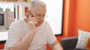 Orta yaşlı, kır saçlı, akıllı telefonlu bir adam yemek odasında masada oturuyor.
