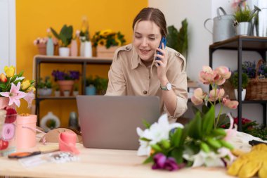 Young caucasian woman florist talking on smartphone using laptop at flower shop